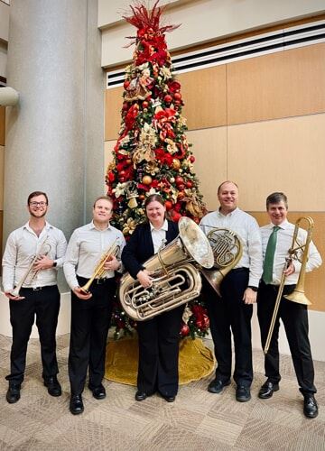 Five musicians pose with their instruments in front of a tall Christmas tree.